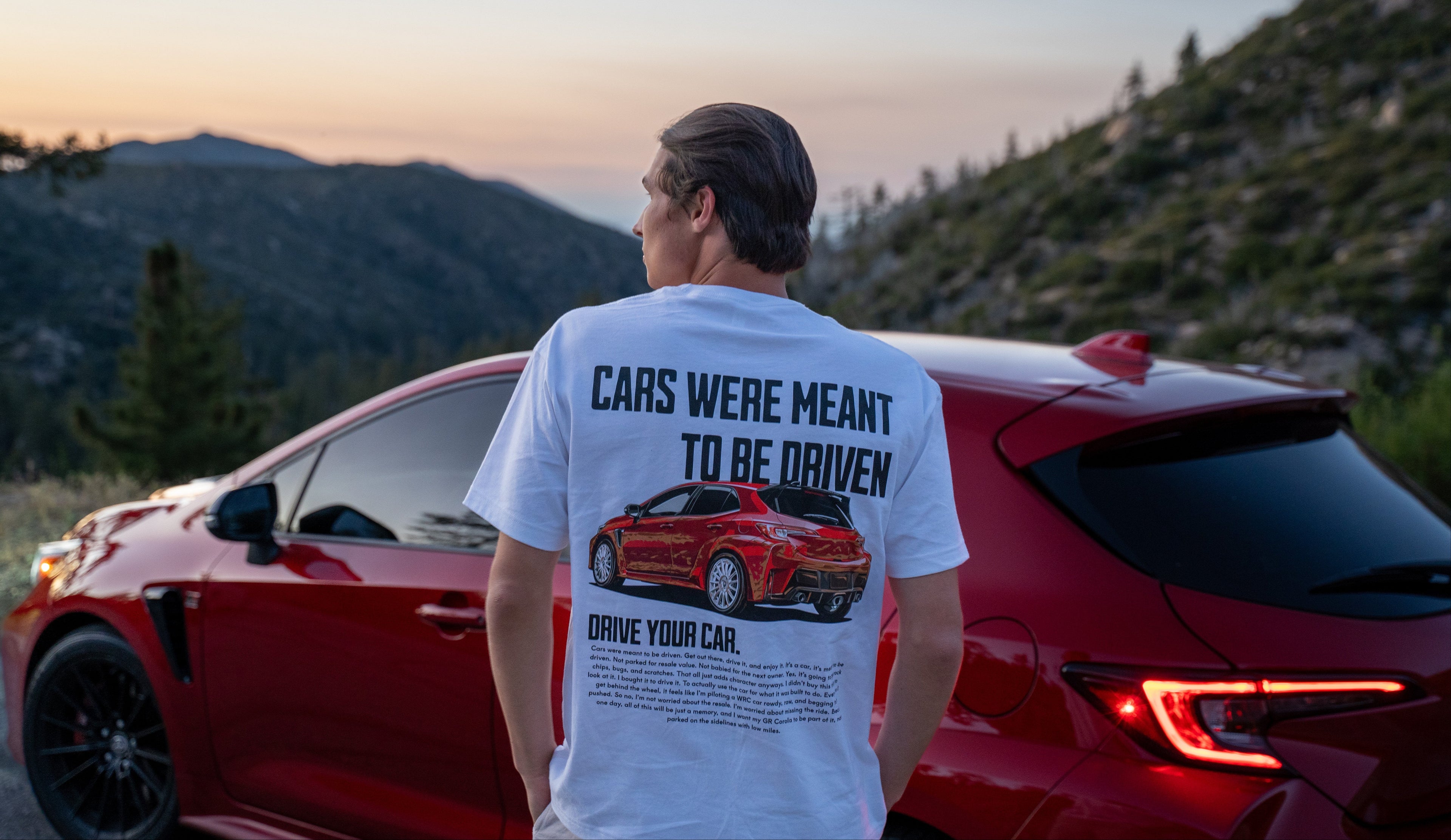 Person wearing a white t-shirt with car-themed text and graphics, standing next to a red car with a mountainous background.